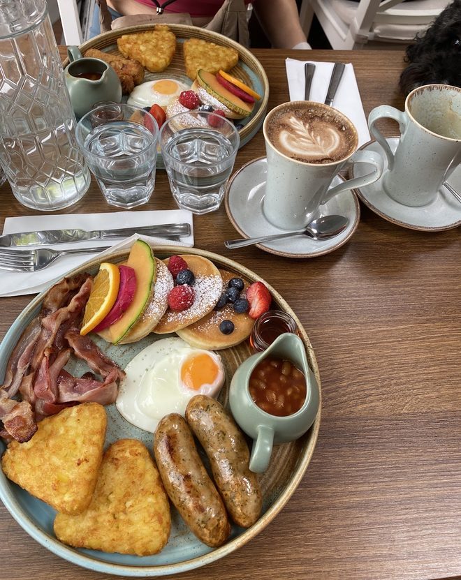 A full English breakfast beside a cappuccino on a wooden table